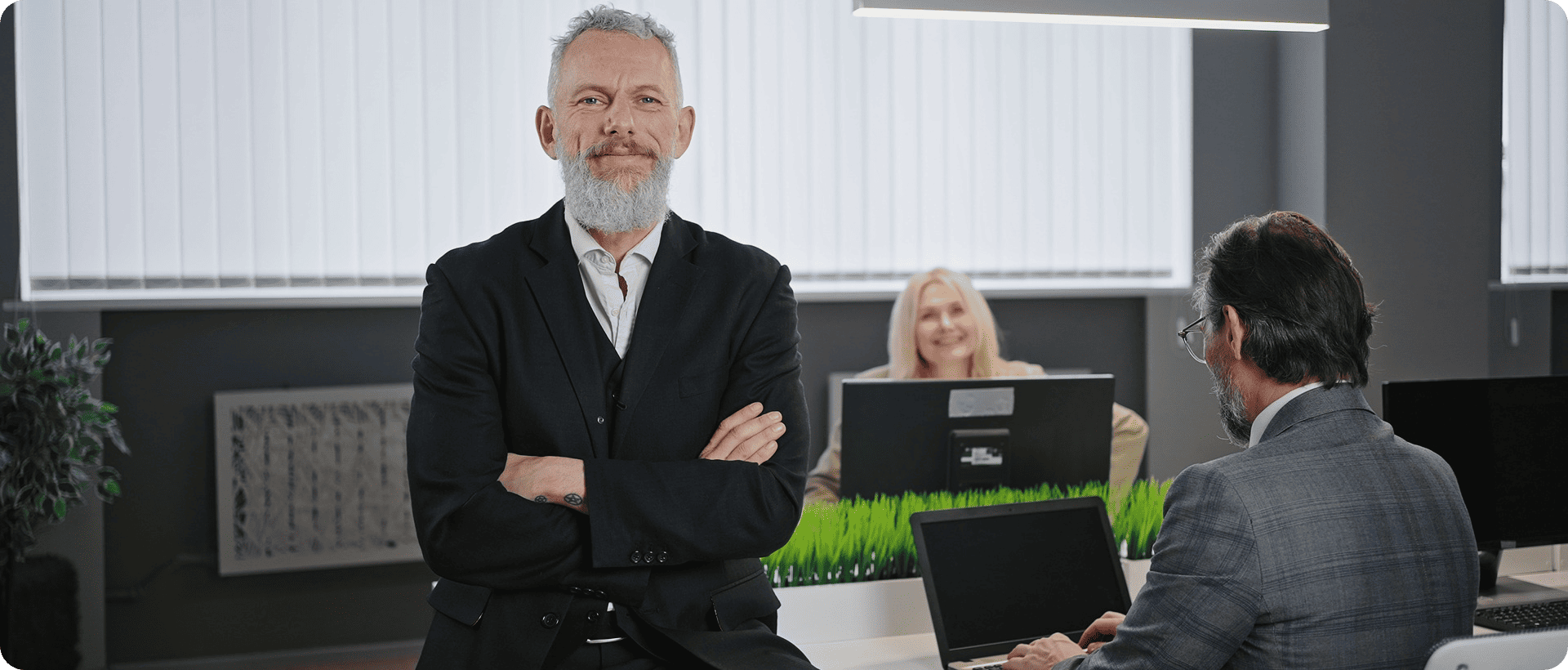 Senior executive standing confidently in a modern office while team members work at computers, illustrating leadership alignment and decision-making speed.